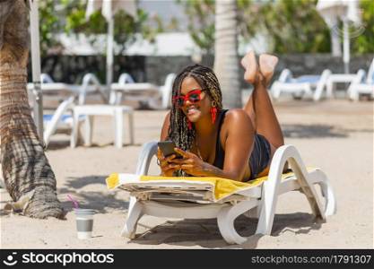 African American woman on the beach lying on a deck chair using phone. Cheerful adult woman enjoying a summer day at the beach.. African American woman on the beach lying on a deck chair using phone