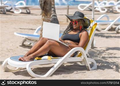African-American woman on the beach relaxing in deck chair using laptop. Cheerful adult woman enjoying a summer day at the beach.. African-American woman on the beach relaxing in deck chair using laptop
