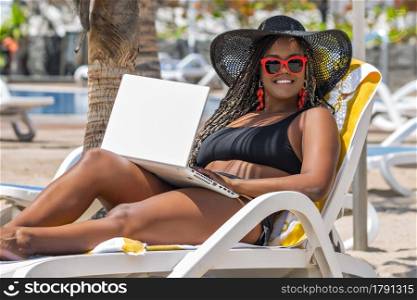 African-American woman on the beach relaxing in deck chair using laptop looking at camera. Cheerful adult woman enjoying a summer day at the beach.. African-American woman on the beach relaxing in deck chair using laptop looking at camera