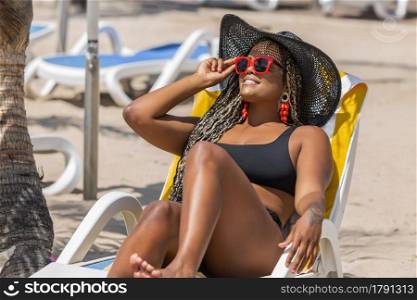 African-American woman on the beach relaxing in deck chair wearing a black hat and holding sunglasses. Cheerful adult woman enjoying a summer day at the beach.. African-American woman on the beach relaxing in deck chair wearing a black hat and holding sunglasses