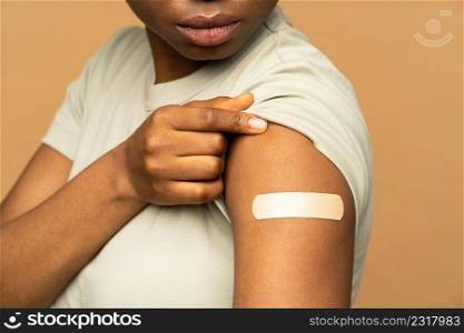 African american woman with adhesive plaster on shoulder after getting coronavirus vaccine isolated on beige wall. Vaccinated black girl and covid-19 vaccination injection. Immunization and health. Young woman look at plaster on shoulder getting coronavirus vaccine. Covid-10 vaccination injection