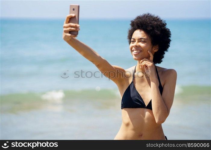 African-American woman with afro hairstyle taking a selfie with her smartphone on a tropical beach. Black girl enjoying her holiday on the beach.. African-American woman with afro hairstyle taking a selfie with her smartphone on a tropical beach.