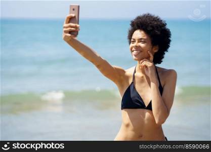 African-American woman with afro hairstyle taking a selfie with her smartphone on a tropical beach. Black girl enjoying her holiday on the beach.. African-American woman with afro hairstyle taking a selfie with her smartphone on a tropical beach.