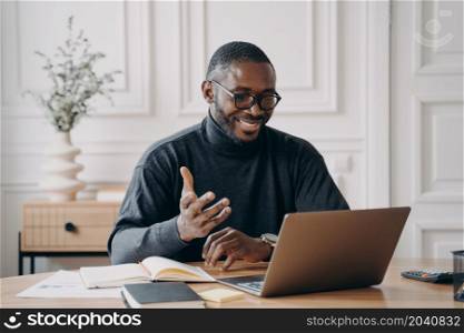 African-American young businessman in glasses having conference online with employes via video call broadly smiling while looking at laptop screen gesturing when communicates and answering questions. African-American young businessman in glasses having conference online with employes via video call