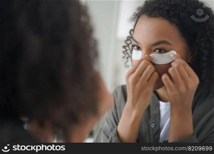 African american young girl apply cosmetic under eye patches, looking at mirror. Pretty mixed race teen lady use under-eye masks to moisturize facial skin. Skincare treatment, morning beauty routine.. African american girl apply cosmetic under eye patches for facial skin. Skincare, beauty routine