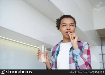 African american young girl taking pill, holding glass of water, pensive biracial woman takes medicine, painkillers or supplements, daily vitamins for skin, natural beauty. Wellness, healthy lifestyle. African american young girl takes pills for skin, natural beauty. Wellness, healthy lifestyle
