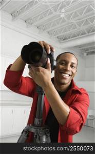 African American young male adult with camera on tripod looking at viewer smiling.