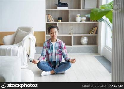 African american young woman is sitting on floor in lotus pose and meditating. Teenage afro girl is practicing yoga at home and exercising lotus position. Spiritual harmony and balance. Stress relief.. Teenage afro girl is practicing yoga at home and exercising lotus position. Stress relief.