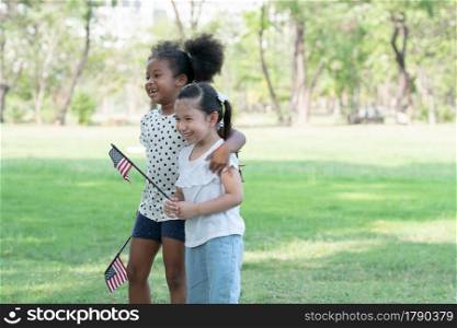 African and Caucasian kids laugh smile hug each other and holding small America flag while children playing in green park. Ethnic diversity of relationship with little girls at 4th of July concept
