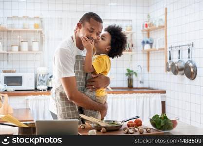 African Cute little boy in yellow casual kissing Dad while cooking at home together. Happy smiling African-American Father while hug and carry his son in kitchen. Joyful Black family, Love emotion.