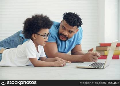 African family father and little son wearing eye glasses lying on floor and online learning with laptop at home
