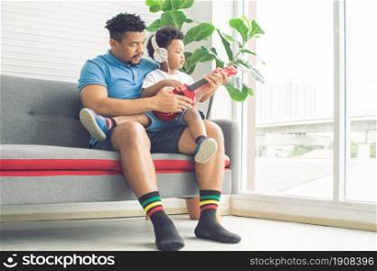 African father and little son are playing ukulele and sitting on sofa in living room at home. Family and Education Concept.