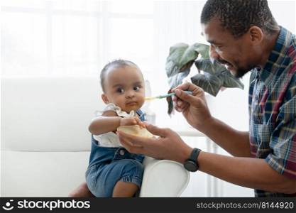 African father feeding adorable toddler baby girl with spoon while sitting on sofa at home and kid"s face mess up with food. Little child care and relationship of dad and little daughter concept