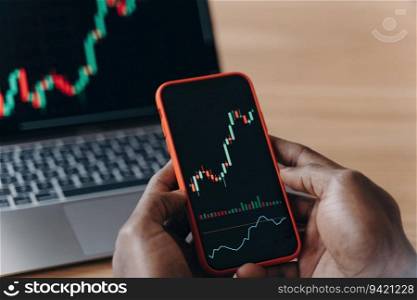 African hands holding smartphone and laptop, analyzing financial data graphs, trading stock market, investment purposes.