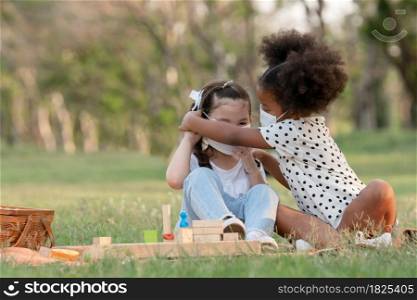 African little girl helping small Caucasian kid wearing face mask while sitting and playing wooden blocks toy in green park. Ethnic diversity of friendship in new normal lifestyle concept