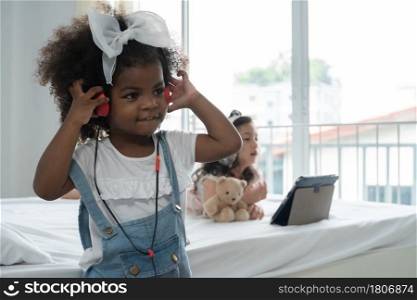 African little kid girl wear headphones enjoy listening music with happy face and her Asian small child friend lying on bed with earphones tablet and hold bear doll at bedroom