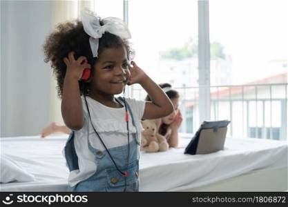African little kid girl wear headphones listening music with happy face while her Asian small child friend lying on bed with earphones tablet and hold bear doll at bedroom