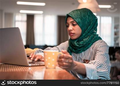 African Muslim businesswoman wearing a green hijab drinking tea while working on laptop computer in relaxation area at modern open plan startup office. High-quality photo. African muslim business woman wearing a green hijab drinking tea while working on laptop computer in relaxation area at modern open plan startup office.