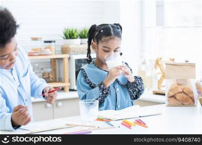 African siblings, little sister drinking milk from glass while brother  playing, drawing together in kitchen at home. Healthy food and drink with children