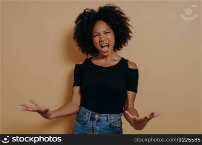 African woman in black t-shirt posing screaming, making palms open, feeling mad, with mouth open wide shouting aloud looking at camera, isolated on beige studio background. Negative emotions concept. African woman in black t-shirt posing screaming, making palms open, feeling mad