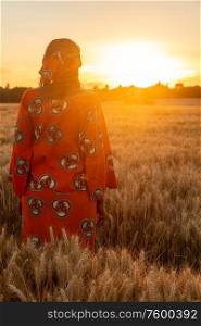 African woman in traditional clothes standing in field of barley or wheat crops at sunset or sunrise