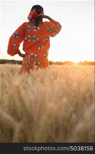 African woman in traditional clothes standing looking across field of barley or wheat crops at sunset or sunrise