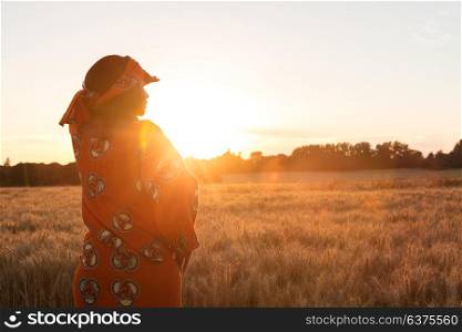 African woman in traditional clothes standing, looking, hand to eyes, in field of barley or wheat crops at sunset or sunrise