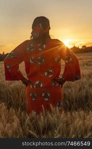 African woman in traditional clothes standing, looking, hands on hips, in field of barley or wheat crops at sunset or sunrise