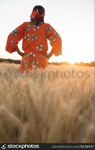 African woman in traditional clothes standing with her hands on her hips in field of barley or wheat crops at sunset or sunrise