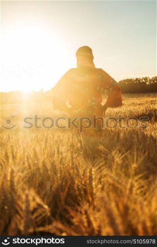 African woman in traditional clothes standing with her hands on her hips in field of barley or wheat crops at sunset or sunrise