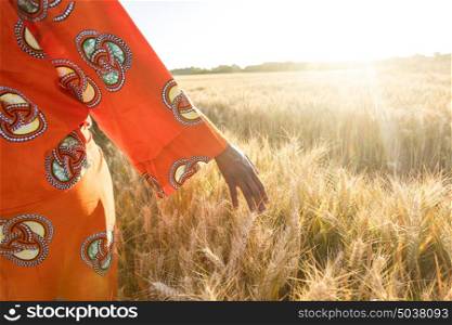 African woman in traditional clothes walking with her hand touching field of barley or wheat crops at sunset or sunrise