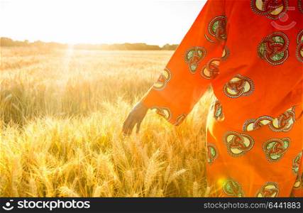 African woman in traditional clothes walking with her hand touching field of barley or wheat crops at sunset or sunrise