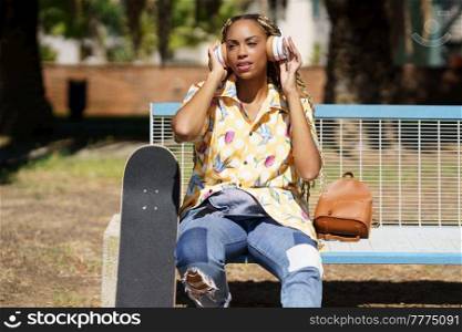 African woman with skateboard. Mixed-race girl relaxing after riding skateboard listening to the music outdoors.. African girl with skateboard relaxing after riding skateboard listening to the music