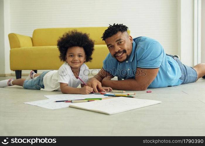African young father and little son smile at camera and lying on warm floor and drawing coloring on book with color pencil crayon at living room