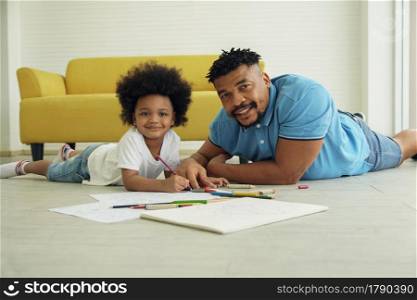 African young father and little son smile at camera and lying on warm floor and drawing coloring on book with color pencil crayon at living room