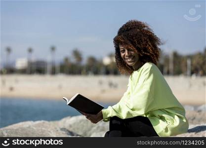 afro american woman sitting on shore while reading a book in a sunny day