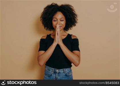 Afro American young woman praying and holding hands in prayer gesture standing isolated on dark beige studio background with copy space, smiling with eyes closed, Praying and hope concept. Afro American young woman praying and holding hands in prayer gesture standing in studio