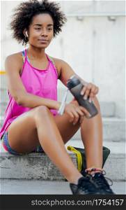 Afro athletic woman drinking water and relaxing after work out while sitting on stairs outdoors. Sport and healthy lifestyle.