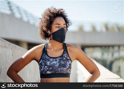Afro athletic woman wearing face mask and relaxing after work out outdoors on the street. New normal lifestyle. Sport and healthy lifestyle.
