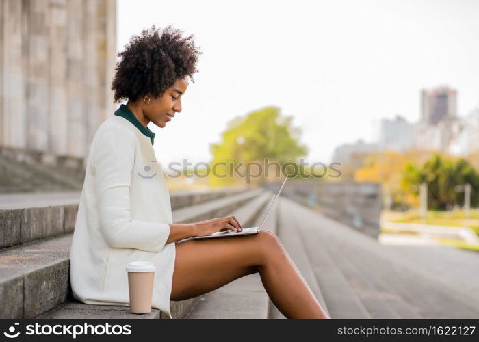 Afro business woman using her laptop while sitting on stairs outdoors. Urban and business concept.