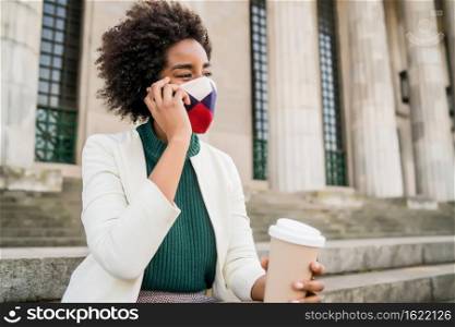 Afro business woman wearing protective mask and talking on the phone while sitting on stairs outdoors at the street. Business and urban concept.