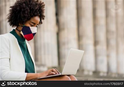 Afro business woman wearing protective mask and using her laptop while sitting on stairs outdoors. Urban and business concept.
