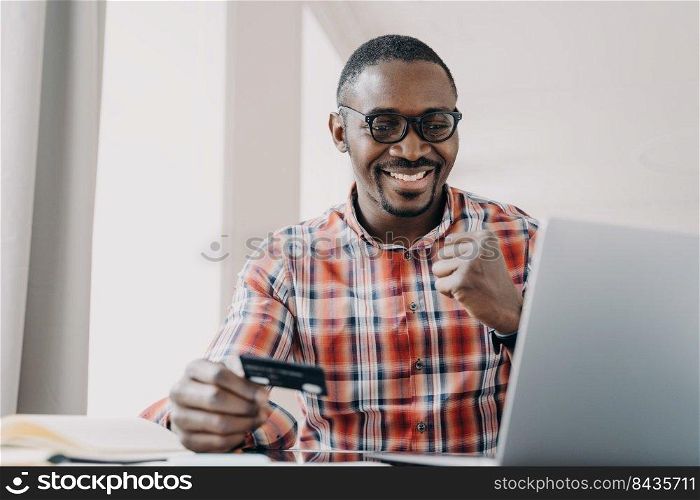 Afro guy gets profit. Happy african american man is buying or ordering online with pc. Businessman has transaction using credit card from home. Ecommerce and consumerism concept. Internet banking.. Afro guy gets profit. Man is buying or ordering online. Ecommerce and consumerism concept.