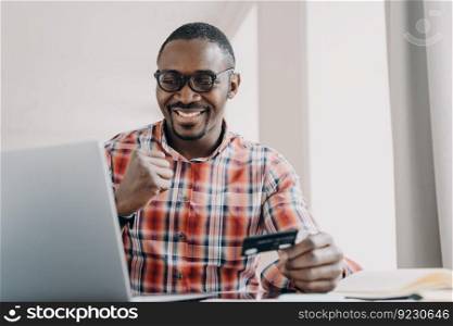 Afro guy gets profit. Happy african american man is buying or ordering online with pc. Businessman has transaction using credit card from home. Ecommerce and consumerism concept. Internet banking.. Afro guy gets profit. Man is buying or ordering online. Ecommerce and consumerism concept.