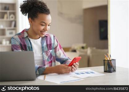 Afro teenage girl is distracting while studying remote. Teenage girl sitting in front of laptop, clicks mobile phone and chatting. African american student gets remote education at school or college.. Afro teenage girl is distracting while studying remote. Student clicks mobile phone and chatting.