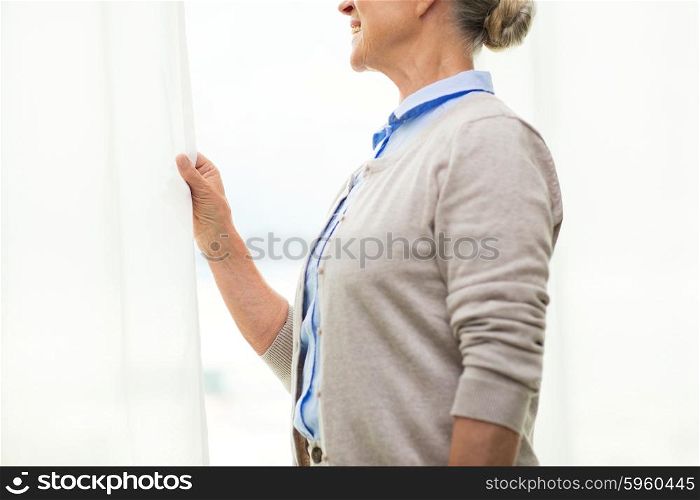 age and people concept - close up of happy smiling senior woman looking through window at home. happy senior woman looking through window at home