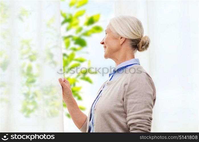 age and people concept - happy smiling senior woman looking through window at home over green natural background. happy senior woman looking through window at home