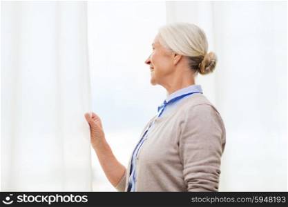 age and people concept - happy smiling senior woman looking through window at home. happy senior woman looking through window at home