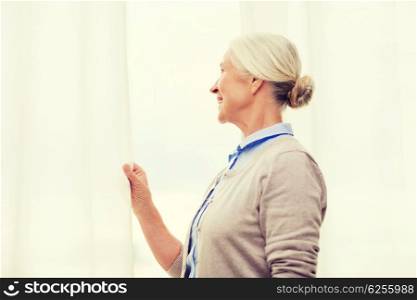age and people concept - happy smiling senior woman looking through window at home. happy senior woman looking through window at home
