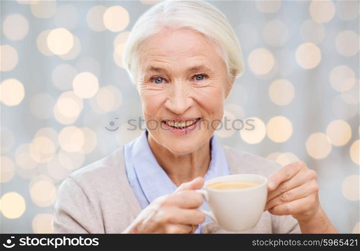 age, drink and people concept - happy smiling senior woman with cup of coffee over holidays lights background